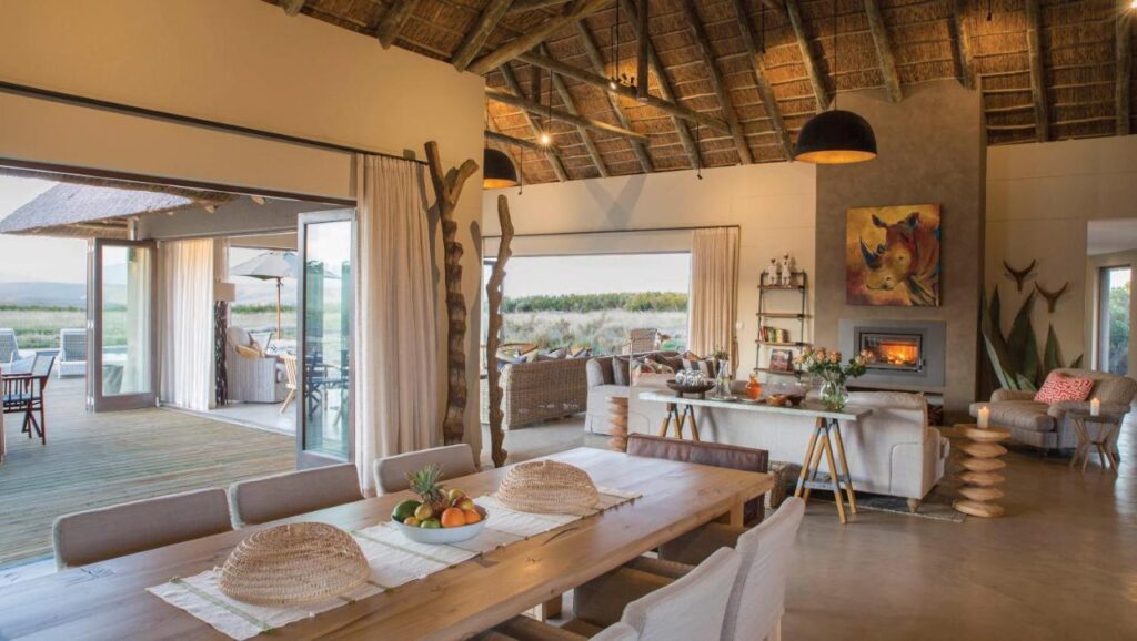 Open-plan living and dining area with wooden table, beige sofas, thatched roof, fireplace, animal-themed artwork—perfect for Game Lodges in Western Cape—with large open doors leading to a patio offering outdoor seating and scenic views.