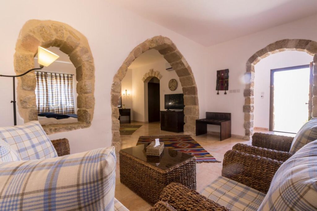Authentic Hotels in Petra Jordan: Stay near the Ancient City 4 A cozy living room with wicker furniture and plaid cushions, reminiscent of boutique hotels in Petra, Jordan, features arched stone doorways, a glass coffee table, and sunlight streaming in through a window and open door.