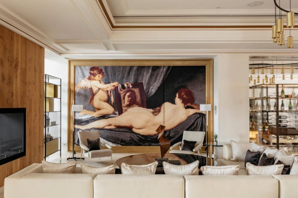 Where to Stay in Madrid: 14 Best Hotels 17 A luxurious living room, ideal for those wondering where to stay in Madrid first time, features a large classical painting of a reclining nude woman and cupid, cream sofas with cushions, a round coffee table, modern decor, and bottles on display.