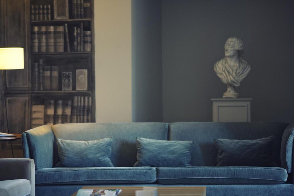 25 Weekend Trips from Madrid Max. 2h Away 91 A cozy living room with a blue sofa and three cushions, a lamp, a wall mural of bookshelves, and a white classical bust on a pedestal—the perfect spot to plan weekend trips from Madrid.