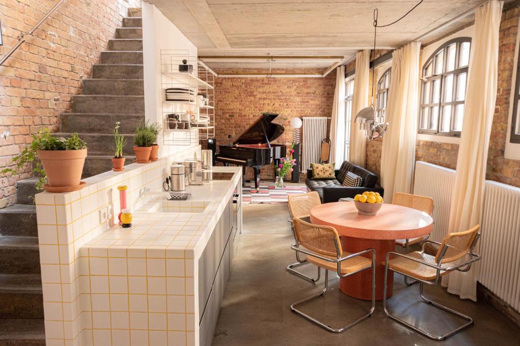 Boutique Hotels in Berlin Kreuzberg 24 Loft-style living space with exposed brick walls, a tiled kitchen island, boutique hotels in Berlin Kreuzberg-inspired dƩcor, potted plants, round dining table with woven chairs, large curtained windows, and a black grand piano near the sofa.