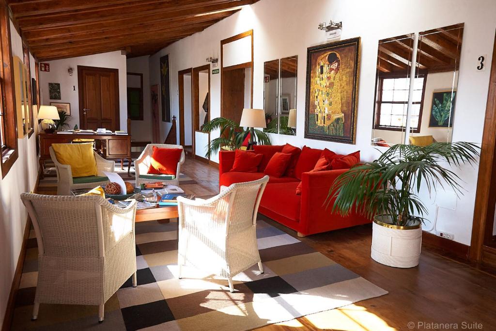 11 UNIQUE Boutique Hotels in Tenerife 26 Bright living room with wooden floors and ceiling, red sofa, white wicker chairs, large mirrors, potted plants, and artwork—like those found in boutique hotels in Tenerife—flooded with natural light and featuring a striped rug by the coffee table.