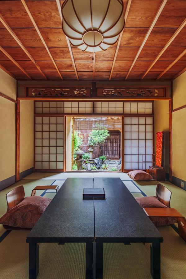 7 UNIQUE Ryokans in Gion Kyoto 9 A traditional Japanese room like those found in ryokans in Gion, Kyoto, with tatami mats, a low black table, floor cushions, shoji sliding doors, and a garden view. Wooden ceiling and soft lighting create a calm, inviting atmosphere.