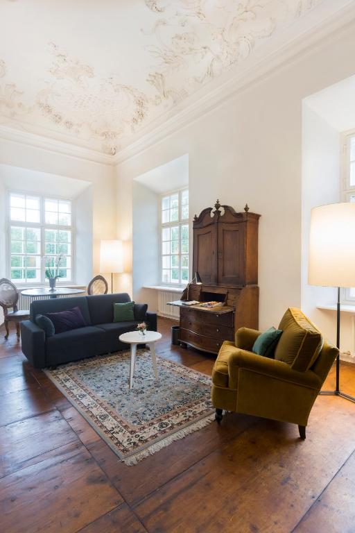 Unique Boutique Hotels in Salzburg 13 A bright living room reminiscent of Boutique Hotels in Salzburg, with large windows, a gray sofa, green armchair, wooden cabinet, white side table, rug, and tall floor lamps on hardwood floors beneath an ornate white ceiling.