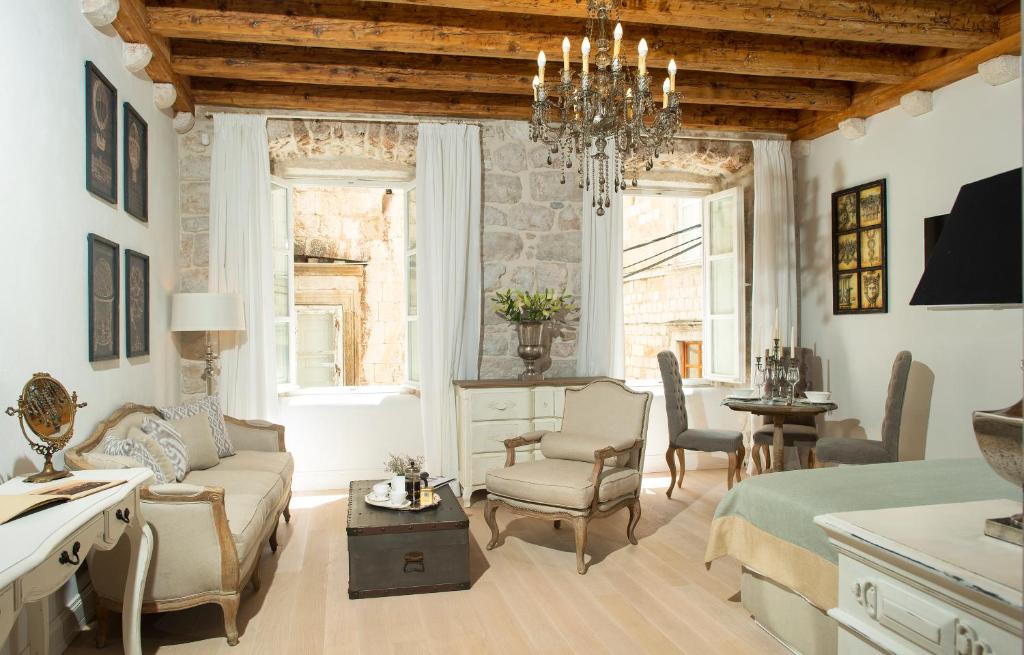 Most Unique Boutique Hotels in Dubrovnik 33 Elegant, rustic living room reminiscent of boutique hotels in Dubrovnik, with exposed wood ceiling beams, stone walls, antique-style furniture, chandelier, and natural light streaming through an open window; cozy sitting and dining areas are visible.