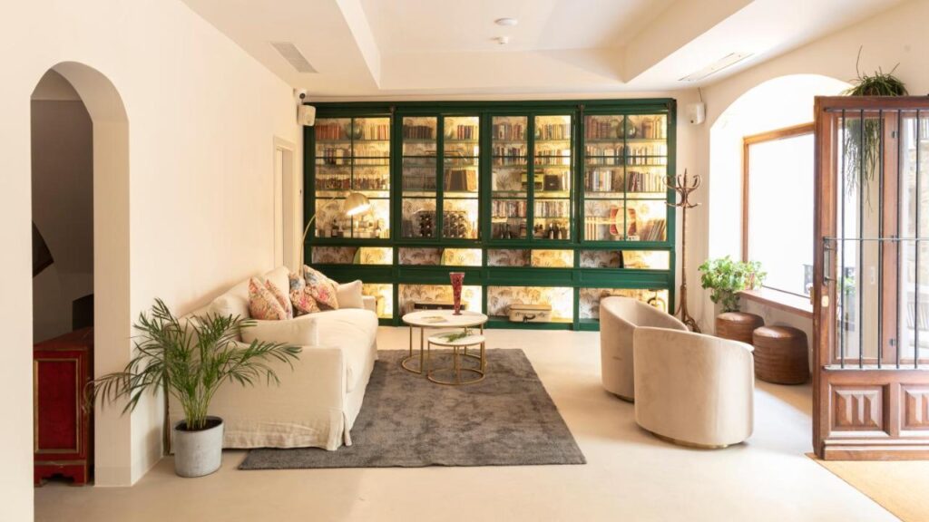 A bright, modern living room reminiscent of boutique hotels in Costa Brava, featuring a white sofa, beige armchairs, a gray rug, round coffee table, lush plants, and a stylish bookshelf with glass doors bathed in sunlight from a large window.