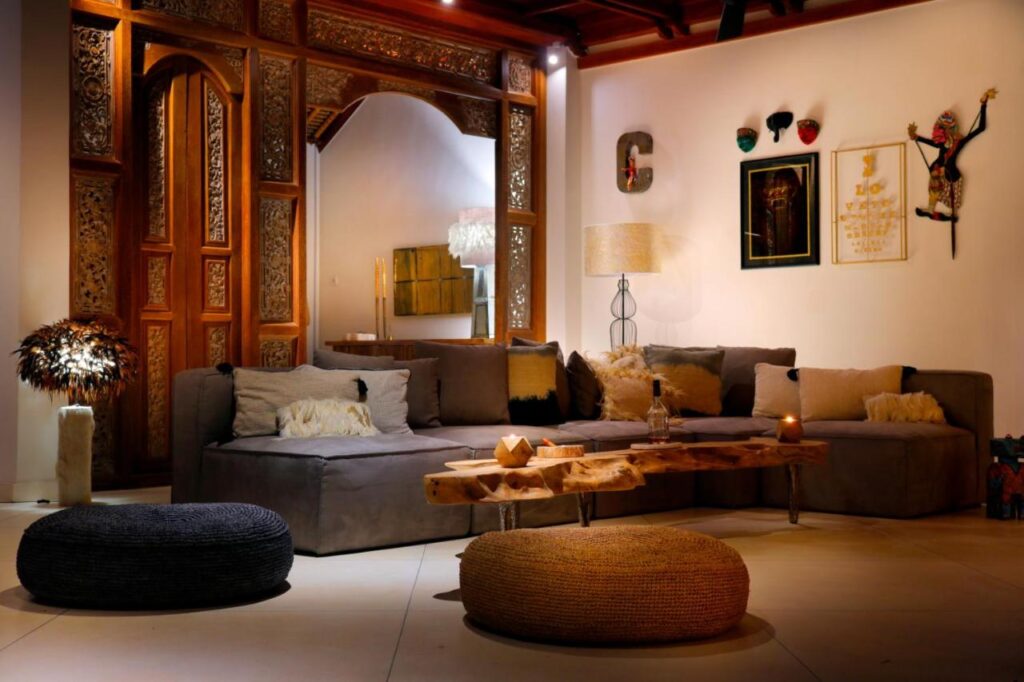 A cozy living room, inspired by the best family villas in Bali, features a gray sectional sofa, fluffy pillows, rustic wooden coffee table, and woven poufs. An ornate wooden arch, wall art, masks, and warm lighting create a welcoming, eclectic atmosphere.