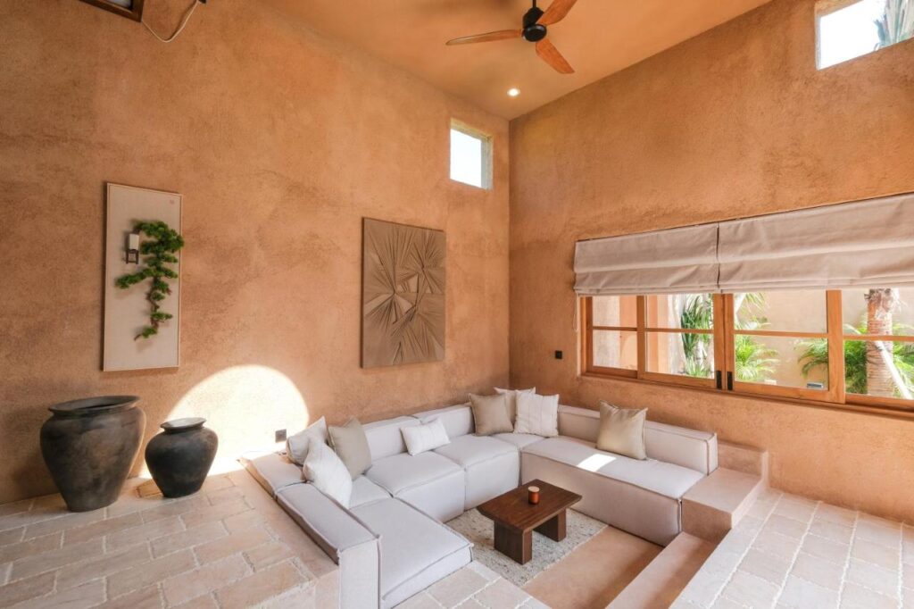 A spacious, sunlit living room in one of the best family villas in Bali, featuring beige walls, a white sectional sofa, a small wooden coffee table, large ceramic vases, wall art, and a window with white curtains letting in natural light.