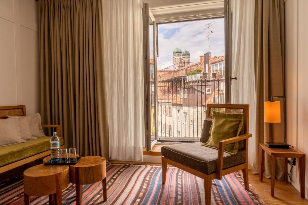 Authentic Boutique Hotels in Munich 13 Sunny room with a chair, side tables, and sofa, featuring a striped rug and open window with sheer curtains. The view recalls boutique hotels in Munich, overlooking city rooftops and a distant church with green domes.