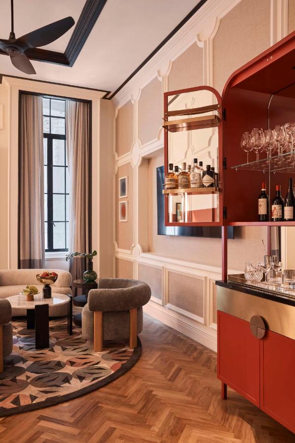 13 Most Unique Hotels in Singapore for an Incredible Holiday 29 A stylish living room inspired by unique hotels in Singapore, with a geometric rug, modern gray armchairs, a round black coffee table, tall windows with beige curtains, and a red wall-mounted bar cabinet displaying bottles and glassware.