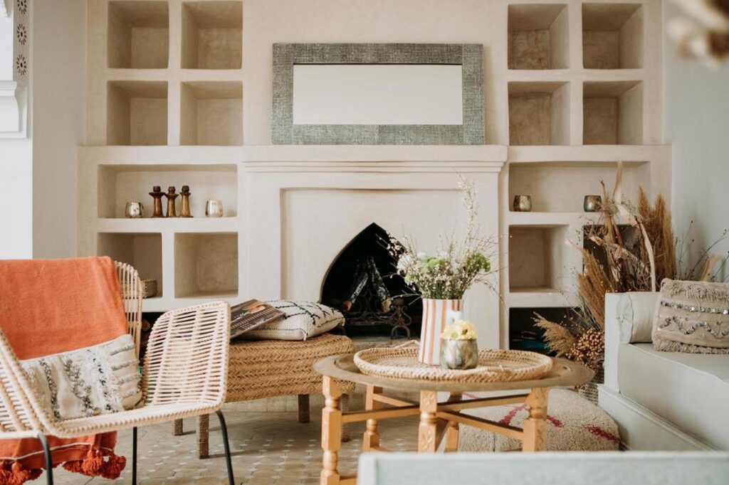 12 Charming Budget Riads in Marrakech 31 A cozy living room, inspired by budget riads in Marrakech, features wicker chairs, a round wooden table, built-in shelves around a fireplace, dried plants, and neutral-toned decor. A rectangular mirror hangs above the fireplace.