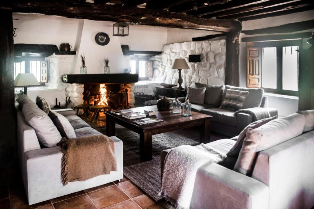 25 Weekend Trips from Madrid Max. 2h Away 67 Cozy rustic living room with a stone fireplace, wooden ceiling beams, and three sofas around a wooden coffee table—perfect for relaxing after weekend trips from Madrid. Soft lighting and large windows create a warm, inviting atmosphere.