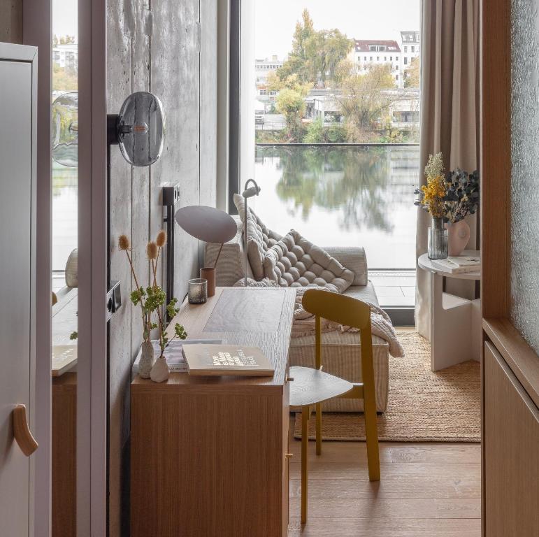 Boutique Hotels in Berlin Kreuzberg 33 A cozy, modern apartment interior reminiscent of boutique hotels in Berlin Kreuzberg, featuring a small desk, wooden chair, and sofa facing large windows overlooking a calm river and cityscape in warm neutral tones.
