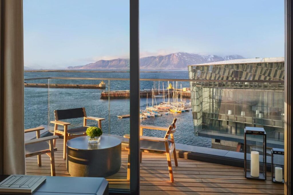 Where to Stay in Reykjavík: Top Guide 3 A modern balcony with wooden chairs and a table overlooks a marina with sailboats, calm blue water, and a contemporary glass building—an ideal spot for those seeking where to stay in Reykjavik, with mountains under a clear sky in the distance.