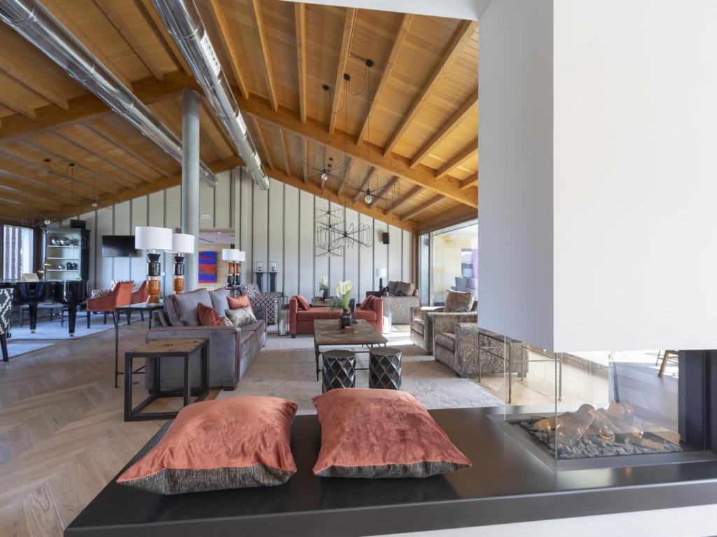 A spacious modern living room, inspired by boutique hotels in Asturias, features a wooden vaulted ceiling, exposed metal ducts, stylish furniture, and a fireplace. Pink cushions sit on a bench in the foreground, with sofas and a dining area behind.