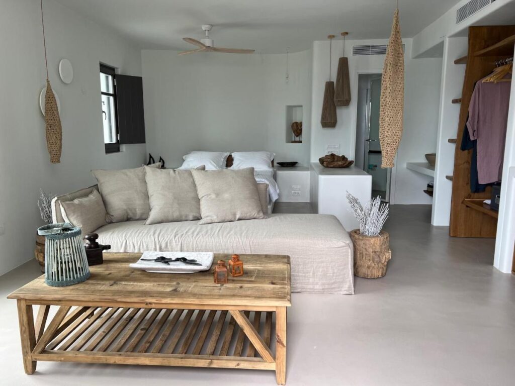 Authentic Hotels in Sikinos, Cyclades 4 A modern, minimalist bedroom with neutral tones, featuring a bed, light sofa, wooden coffee table, and natural decor accents—evoking the serene style of boutique hotels in Sikinos—with an open closet and large windows letting in natural light.
