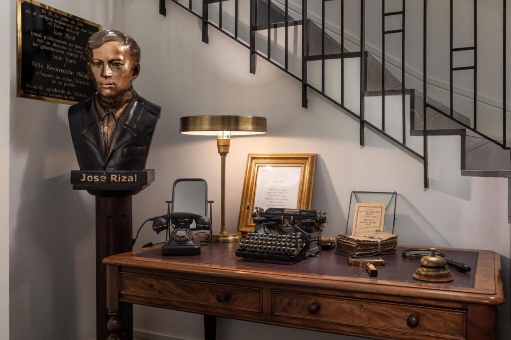 Where to Stay in Madrid: 14 Best Hotels 14 A wooden desk under a staircase holds a vintage typewriter, rotary phone, framed documents, books, a lamp, and a gavel—an inspiring scene reminiscent of discovering where to stay in Madrid first time. A bust of José Rizal sits to the left with a plaque behind it.