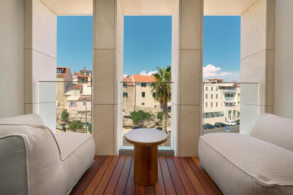 8 TOP Boutique Hotels in Split 22 Two white lounge chairs and a small round wooden table sit on a wooden floor by tall windows overlooking old stone buildings, palm trees, and a bright blue sky—a perfect spot for those seeking where to stay in Split.