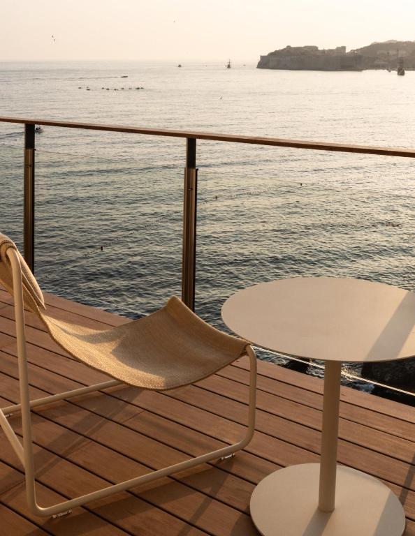 Most Unique Boutique Hotels in Dubrovnik 2 A beige lounge chair and a small round table sit on a wooden deck overlooking calm sea water, echoing the serene ambiance found at Boutique Hotels in Dubrovnik, with a glass railing and hazy coastline under soft sunlight.