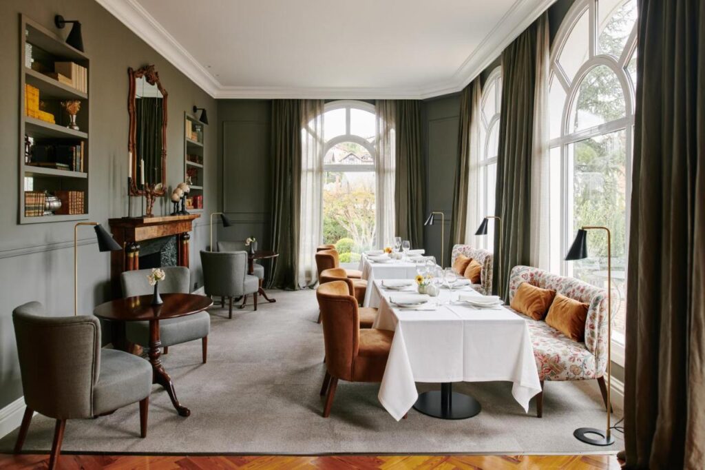 Best Boutique Hotels in San Sebastian 15 Elegant dining room with large arched windows, tall curtains, grey walls, and a mix of armchairs and sofas around tables set with white linens—reminiscent of boutique hotels in San Sebastian. Warm, cozy, and sophisticated atmosphere overlooking a garden.