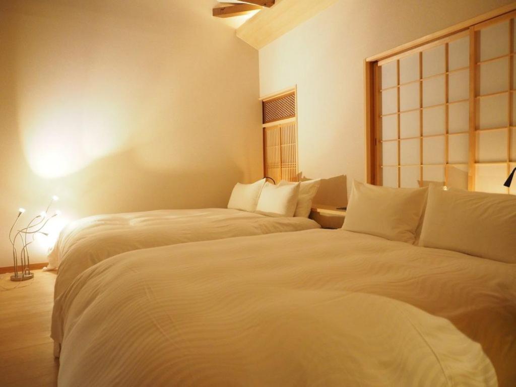 7 UNIQUE Ryokans in Gion Kyoto 4 A serene bedroom inspired by ryokans in Gion Kyoto, with two large beds covered in white linens, soft warm lighting, wooden accents, and a shoji screen on the right wall. A modern floor lamp glows in the back corner.