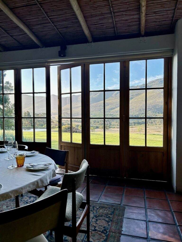 A cozy dining area with set tables and chairs faces large glass doors, revealing a scenic view of green fields and distant mountains under a blue sky—perfect for relaxing at Game Lodges in Western Cape as sunlight streams into the room.