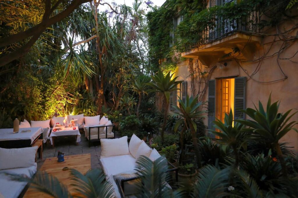 9 UNIQUE Riads in Tangier 5 A cozy outdoor patio with cushioned seating surrounds a fire pit, lush green plants and trees create a serene atmosphere reminiscent of riads in Tangier, while warm light glows from windows of a vine-covered house in the evening.