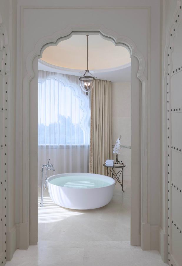 Where to Stay in Doha, Qatar: Transit, City & Beach Hotels 5 A luxurious bathroom—perfect for those seeking where to stay in Doha—features a round white bathtub beneath a large window with sheer curtains, an ornate arched doorway, a pendant light, and a side table with towels and an orchid.