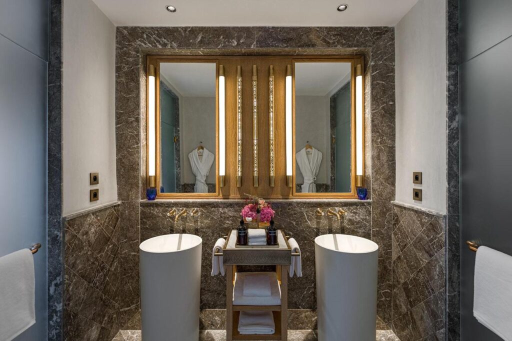 9 UNIQUE Riads in Tangier 9 Modern bathroom with two white pedestal sinks, a marble countertop, and large mirrors—reminiscent of the elegance found in riads in Tangier. Marble walls, plush towels, and a tray of amenities add to the luxurious atmosphere.