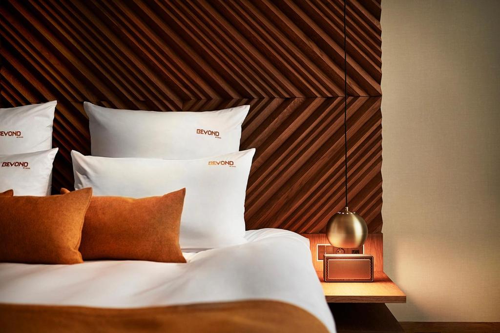 Authentic Boutique Hotels in Munich 35 A modern hotel bed with white linens and brown accent pillows sits beside a wooden nightstand with a round gold lamp—hallmarks of the chic design often found in boutique hotels in Munich. The headboard features a geometric wooden pattern.