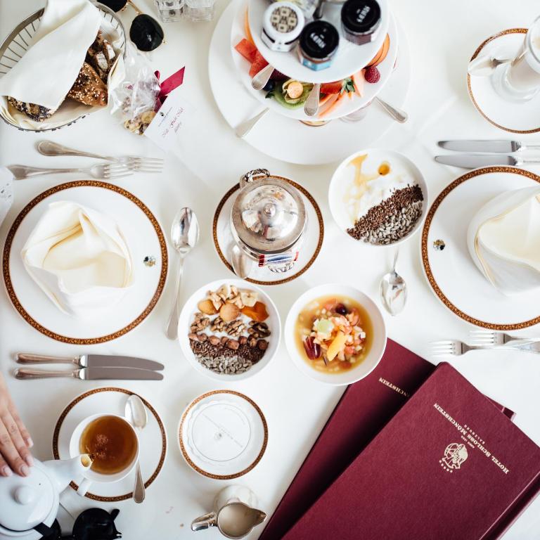 Unique Boutique Hotels in Salzburg 24 A breakfast table at one of the chic Boutique Hotels in Salzburg is set with elegant white and gold dishes, pastries, fruit, granola bowls, tea, a teapot, and maroon menus—captured from above as a hand pours tea in the lower left corner.