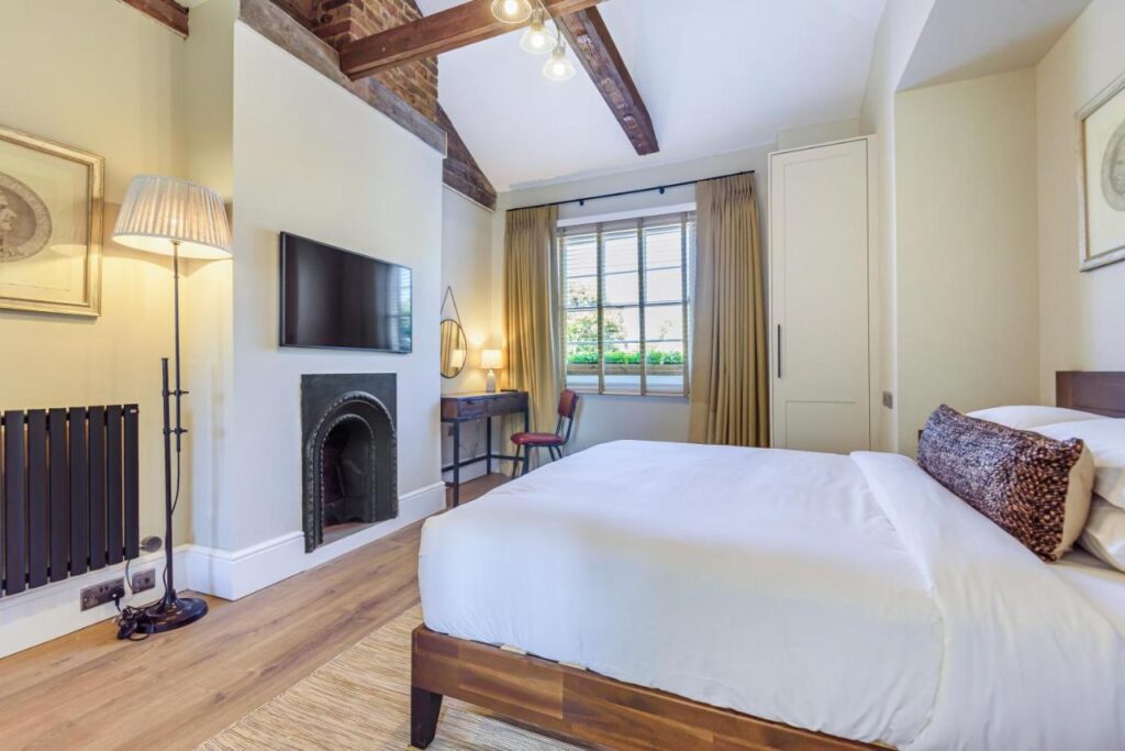 15 Best Boutique Hotels in London, UK 22 A bright bedroom at one of the best boutique hotels London offers, featuring a double bed with white linens, fireplace, wall-mounted TV, desk by the window, exposed wooden beams, modern decor, and abundant natural light through beige curtains.