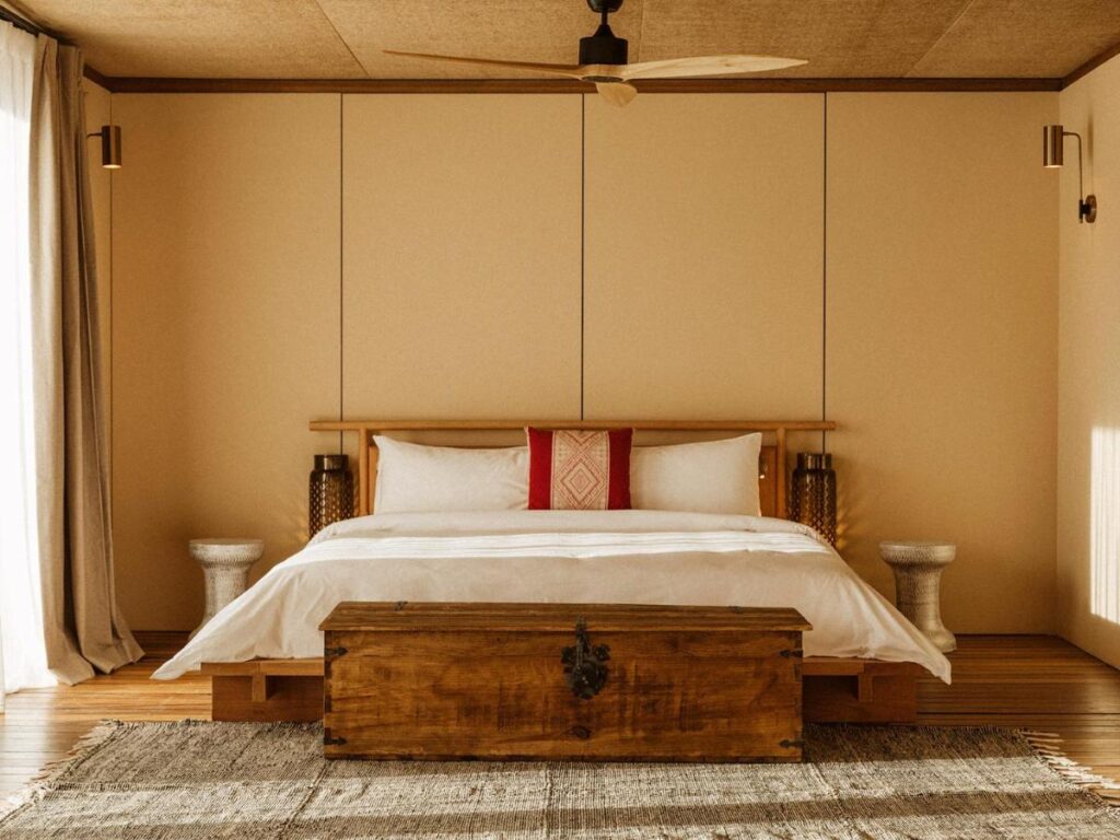 9 Best Hotels in Saudi Arabia: Unique Stays Across the Country 10 Minimalist bedroom inspired by the Best Hotels in Saudi Arabia, featuring a wooden bed frame, white bedding, red accent pillows, side tables, a chest at the foot of the bed, beige walls, ceiling fan, and soft light through left curtains.