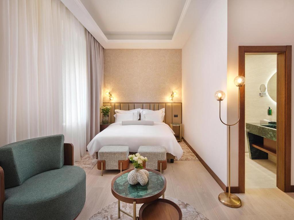 8 TOP Boutique Hotels in Split 28 A modern hotel room with a double bed, round armchair, small coffee table, floor lamp, and en-suite bathroom. The neutral decor and soft lighting evoke the charm of boutique hotels in Split, with sheer curtains on a large window.