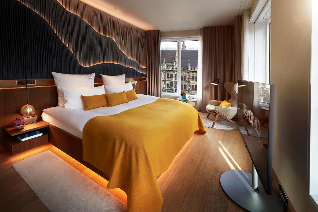 Authentic Boutique Hotels in Munich 32 A modern hotel room in one of the boutique hotels in Munich, featuring a large bed with white linens and a mustard yellow blanket, a TV, reading chair by the window, stylish lighting on a dark accent wall, and city views through large windows.