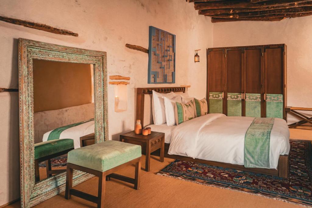 9 Best Hotels in Saudi Arabia: Unique Stays Across the Country 19 A cozy bedroom, reminiscent of the Best Hotels in Saudi Arabia, with a wooden bed, white and green bedding, a large ornate mirror, padded green bench, wooden side table, rug, and wardrobe. The rustic walls feature charming exposed beams.