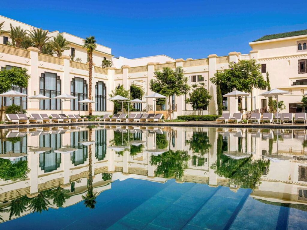 9 UNIQUE Riads in Tangier 6 A luxurious hotel inspired by riads in Tangier features cream-colored walls and green rooftops, reflected in a clear swimming pool. Lounge chairs and white umbrellas line the poolside, surrounded by lush trees under a bright blue sky.