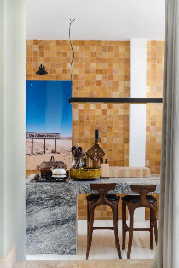 9 UNIQUE Riads in Tangier 25 A modern kitchen with a marble island, two wooden bar stools, yellow tile backsplash, and kitchenware on the counter captures the elegance often found in riads in Tangier. A large blue artwork and exposed light fixture add character to the space.