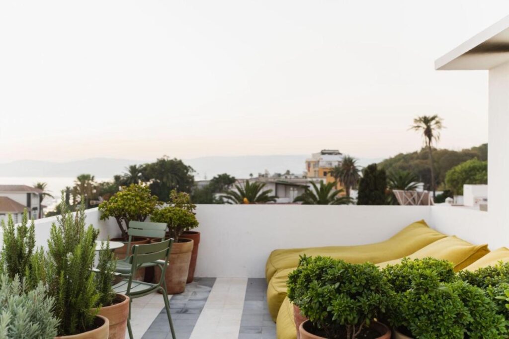9 UNIQUE Riads in Tangier 24 A rooftop terrace with potted plants, a green table and chairs, and yellow lounge beds, reminiscent of stylish riads in Tangier, overlooking buildings, palm trees, and distant hills at sunset.