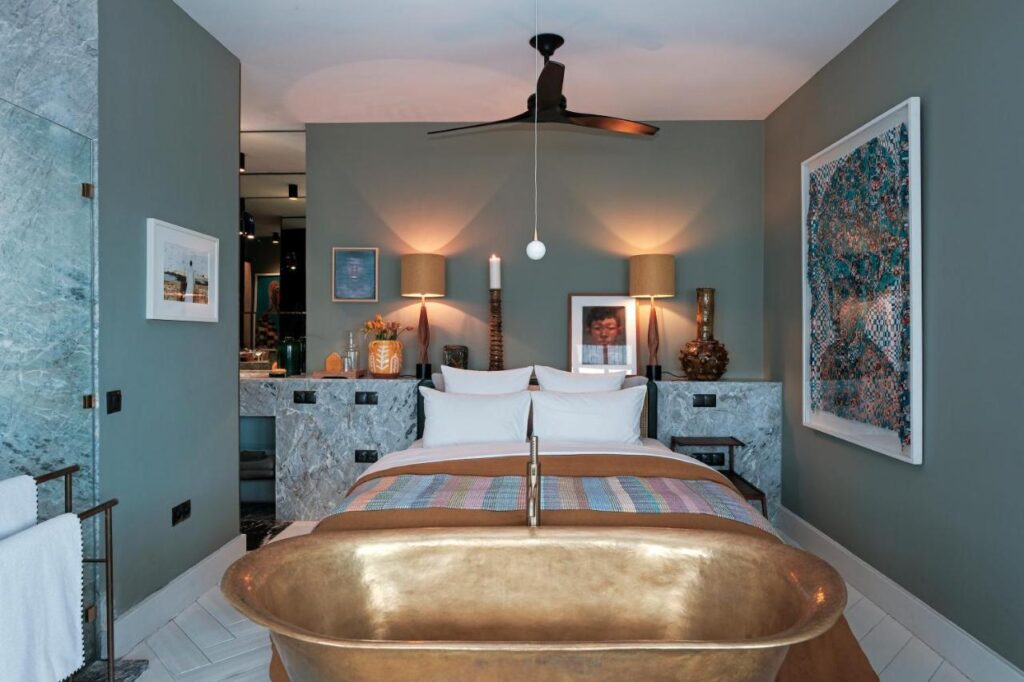 9 UNIQUE Riads in Tangier 22 A stylish bedroom inspired by riads in Tangier, featuring a copper bathtub at the foot of the bed, two bedside tables with matching lamps, wall art, a ceiling fan, and a marble-accented bathroom area in the background.