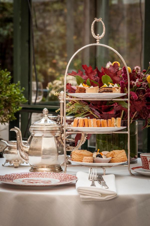 Where to Stay in Madrid: 14 Best Hotels 50 A table set for afternoon tea with a silver teapot, patterned china, and a two-tiered tray of pastries and cakes—an inviting scene reminiscent of discovering where to stay in Madrid first time, elegant and welcoming. Red flowers decorate the background.