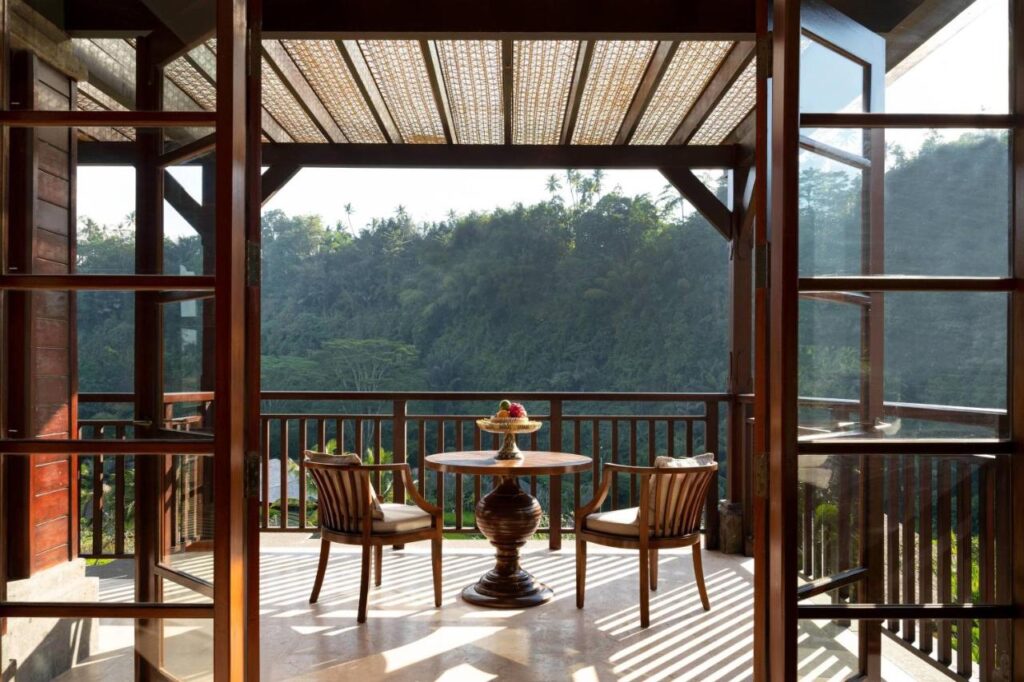 13 Most UNIQUE Places to Stay in Ubud 46 A sunlit balcony with two wooden chairs and a round table overlooks lush green trees and forested hills—open glass doors and a wooden pergola above complete this tranquil retreat among unique places to stay in Ubud.