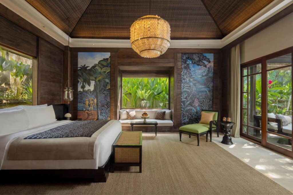 13 Most UNIQUE Places to Stay in Ubud 48 Spacious tropical-themed bedroom with a large bed, woven rug, glass doors, cushioned window seat, green plants outside, mural walls, and a chandelier hanging from a high wood-paneled ceiling—one of the unique places to stay in Ubud.
