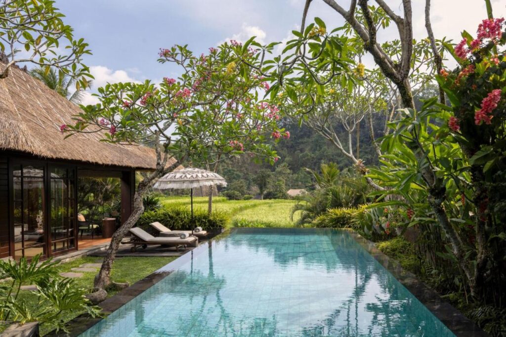 13 Most UNIQUE Places to Stay in Ubud 47 A serene tropical villa with a private pool surrounded by lush greenery and flowering trees, this thatched-roof retreat offers sun loungers and distant hill views—one of the most unique places to stay in Ubud under a partly cloudy sky.