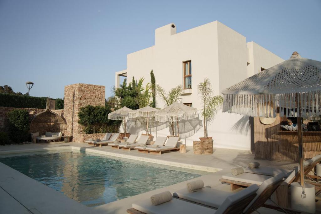 A modern villa with white walls features a swimming pool surrounded by lounge chairs and fringed umbrellas, reminiscent of boutique hotels in Formentera. Lush greenery and a cozy seating area complete the serene outdoor space under a clear sky.