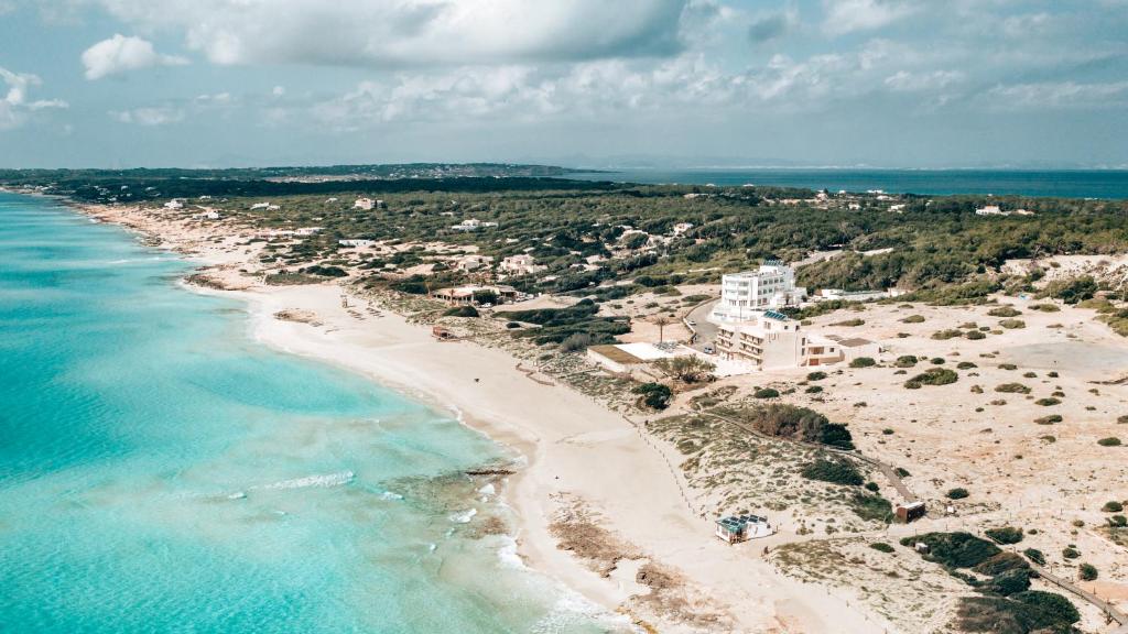 Aerial view of a sandy beach with turquoise water, boutique hotels in Formentera nestled near the shore, and greenery scattered along the coastline under a partly cloudy sky.