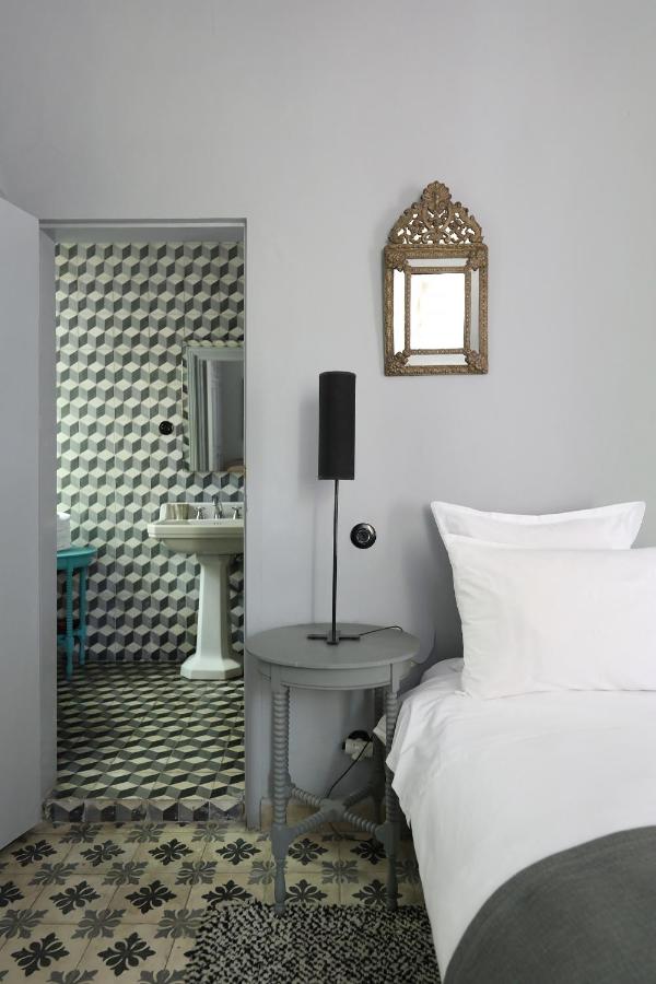 9 UNIQUE Riads in Tangier 3 A modern bedroom inspired by riads in Tangier features a round gray side table, black lamp, and a bed with white linens. An ornate mirror hangs on the light gray wall, while the adjacent bathroom boasts geometric tiles and a white pedestal sink.