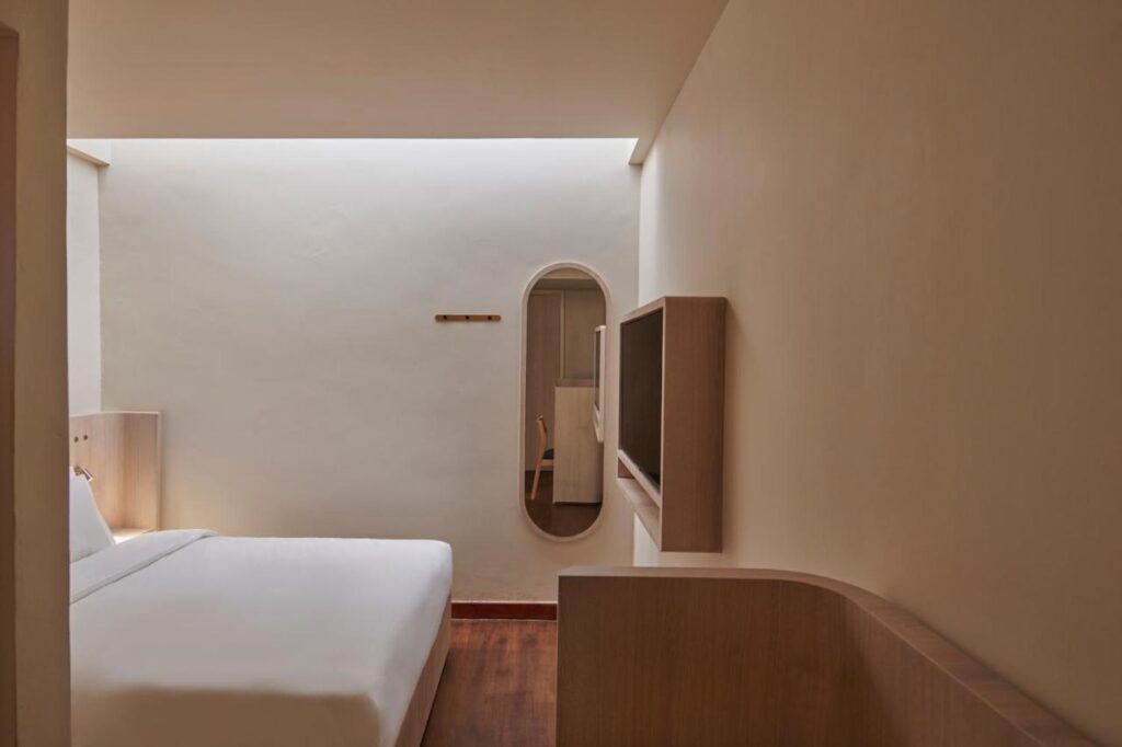 13 Most Unique Hotels in Singapore for an Incredible Holiday 10 Minimalist hotel room with a double bed, white bedding, wooden floor, wall-mounted TV, oval mirror, and simple coat rack on a plain beige wall. Soft natural light from above creates a calm retreat among unique hotels in Singapore.