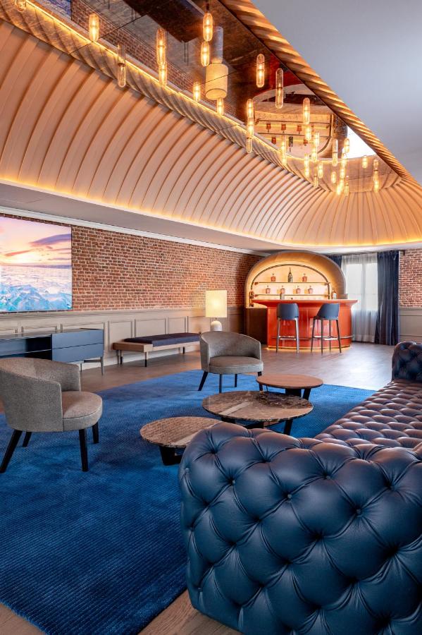 Where to Stay in Madrid: 14 Best Hotels 58 Modern lounge with tufted blue sofa, round tables, armchairs, bar stools, brick walls, large TV, and mirrored ceiling lights. Blue carpet covers the floor as natural light enters—ideal for those wondering where to stay in Madrid first time.