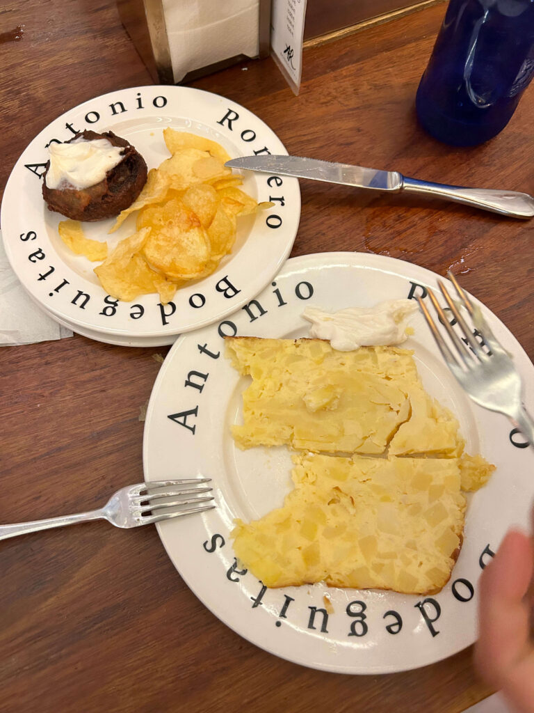 1 day in Seville Spain: My Favorite City 15 Two plates on a wooden table: one with potato chips and a meatball topped with sauce, the other with Spanish omelette slices and aioli—perfect bites to enjoy during 1 day in Seville, Spain. Two forks rest nearby, ready for sharing.